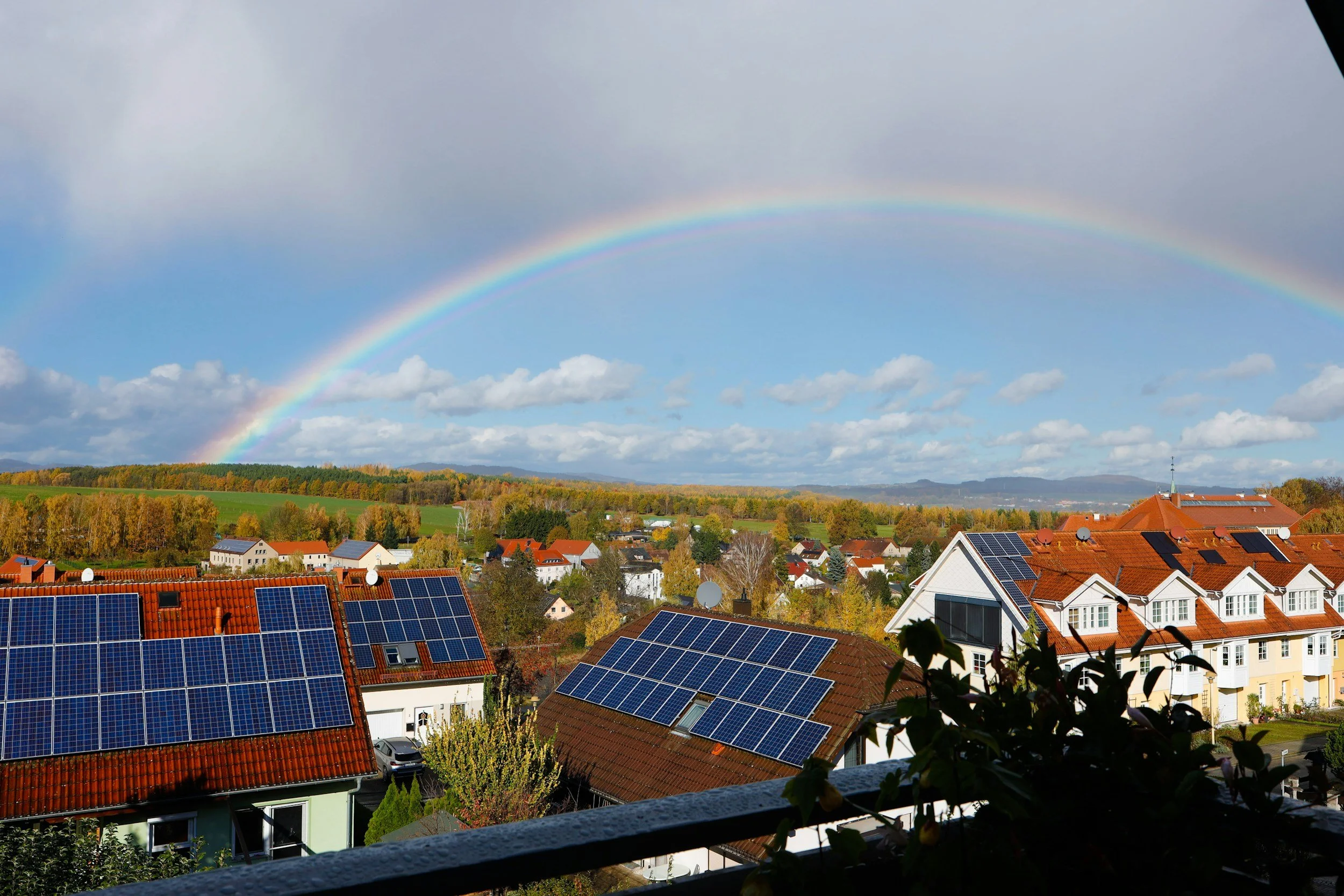 A double rainbow arching across a partly cloudy sky over a residential area with red-tiled roofs and solar panels. There are green fields, trees, and distant hills in the background.