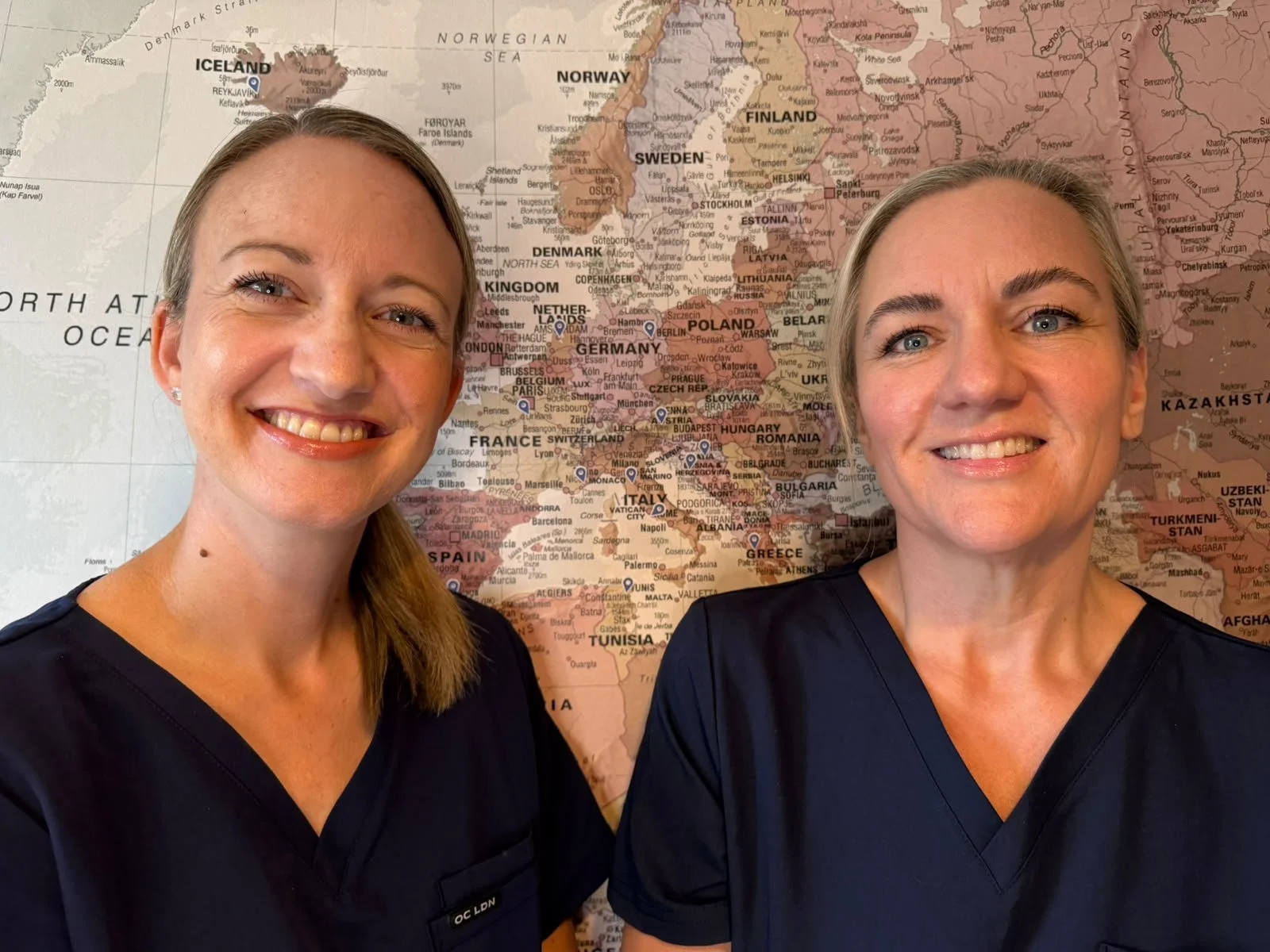 Two women wearing navy scrubs posing in front of a world map, focusing on Europe and northern Africa.