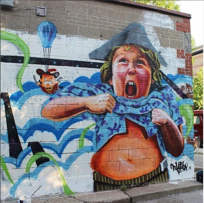 A graffiti mural of a girl with blonde hair, yelling with her mouth wide open, pulling up her shirt to reveal her stomach. The mural is on a brick wall with blue and white clouds, a green balloon, and a cartoon bear in the sky. Art, street art, paint