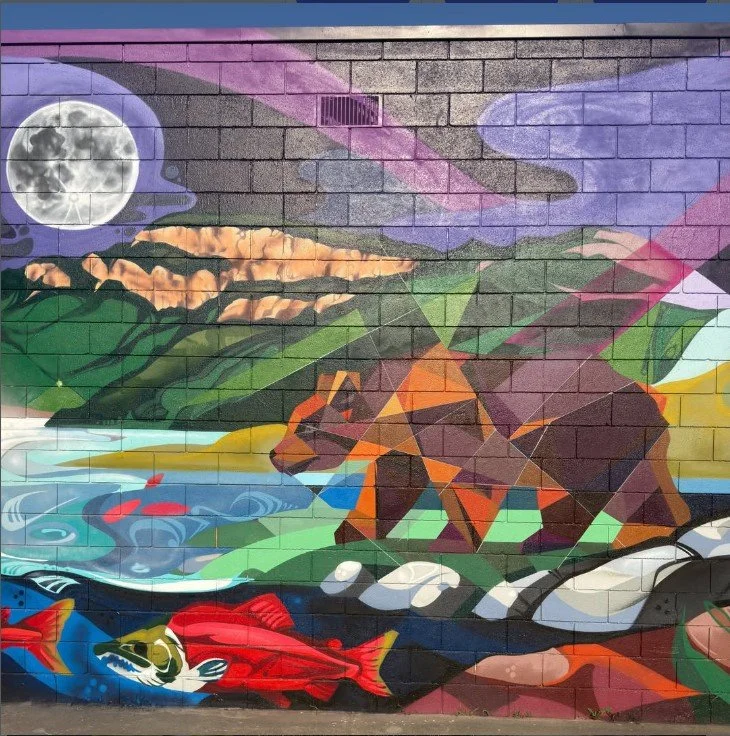 A colorful mural painted on a brick wall depicting a bear, a fish in water, mountains, trees, a full moon, and a purple sky.