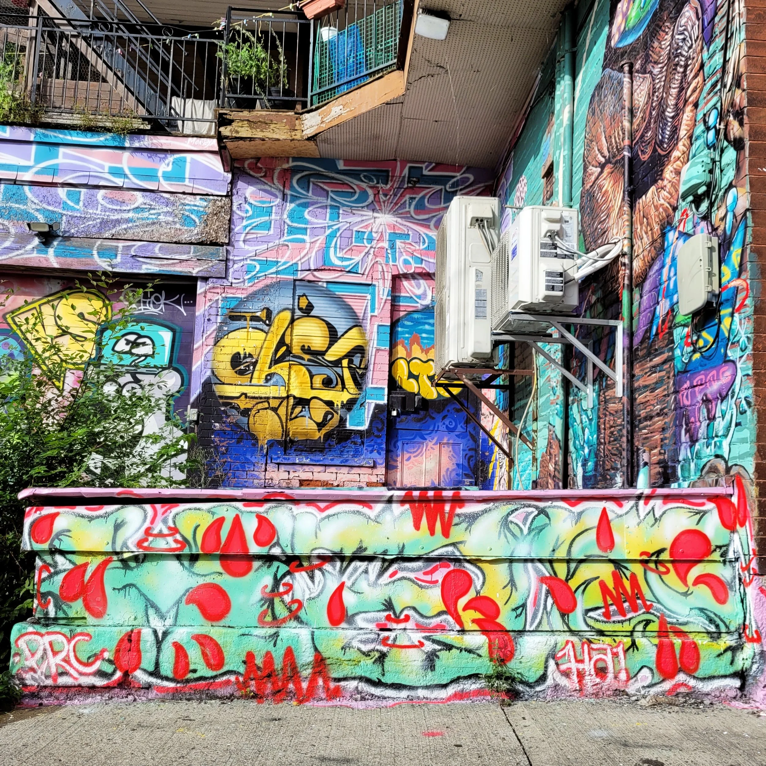 Colorful graffiti covering a brick wall, with various tags, designs, and bright colors. There are air conditioning units attached to the wall, with small plants growing near the ground and some on a nearby balcony.