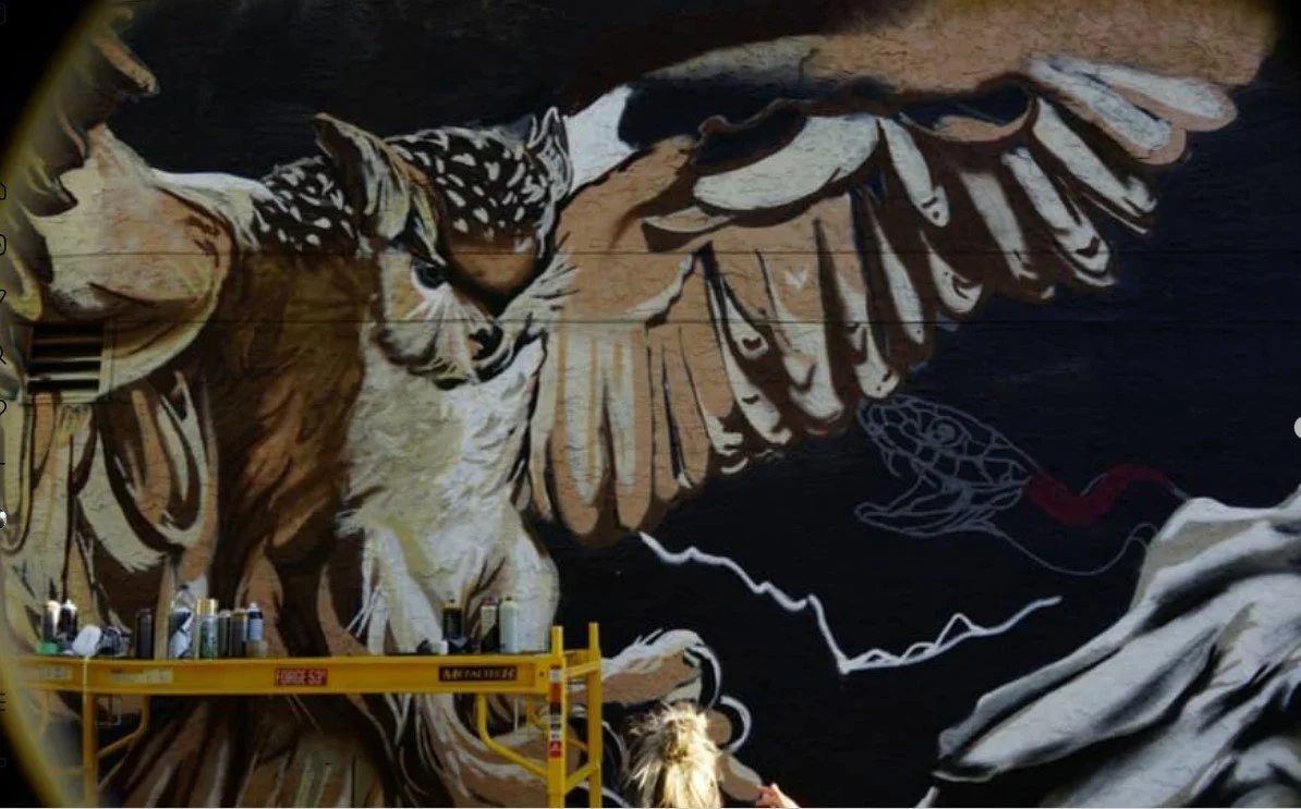 A mural of an eagle with outstretched wings in progress, with an artist working on it from a yellow lift.