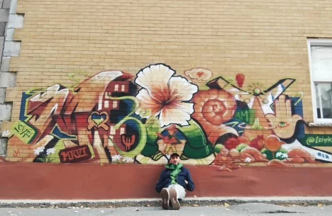A boy sitting in front of a colorful graffiti mural on a wall, featuring various abstract shapes, a shell, and other decorative elements. Art, street art, painter, mural, graffiti, spray paint, oil paint, montreal, commission, Futurism, retro Futuris