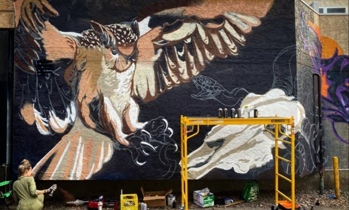 A mural of a large owl with outstretched wings next to a mountain and a small bird, being painted outdoors with scaffolding and painting supplies nearby.