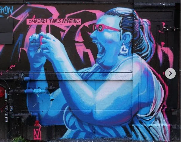 Street art mural of a woman with blue skin, wearing sunglasses and earrings, excitedly looking at her phone with a speech bubble saying "OMG! GAD! THAT'S AMAZING.", Art, street art, painter, mural, graffiti, spray paint, oil paint, montreal, commissi
