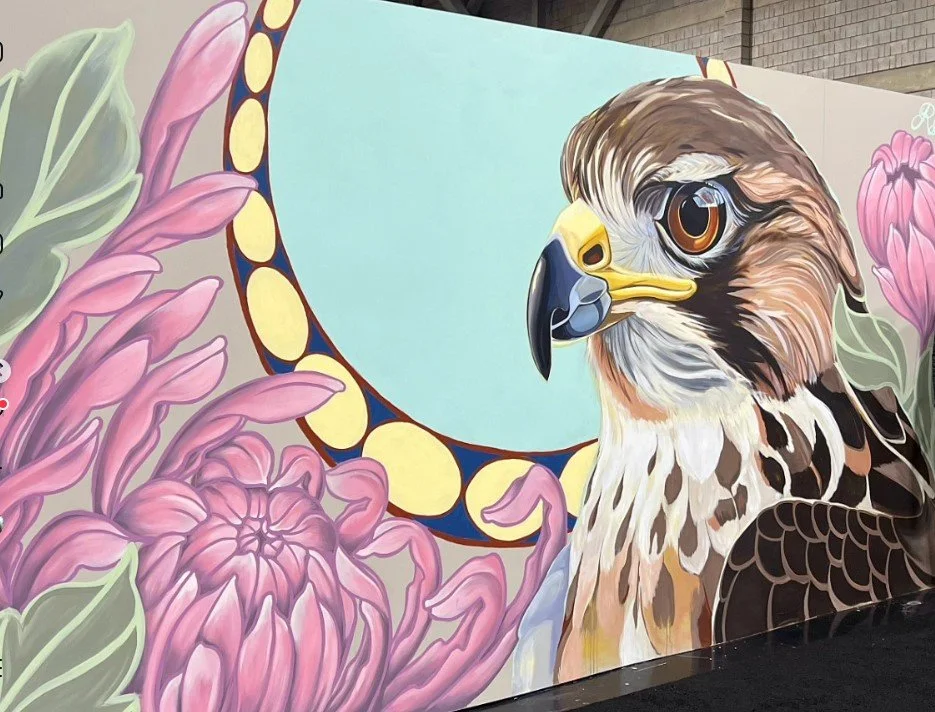 Colorful mural of a bird of prey, likely a hawk or falcon, with large brown eyes, detailed feathers, surrounded by pink flowers and a decorative circular background.