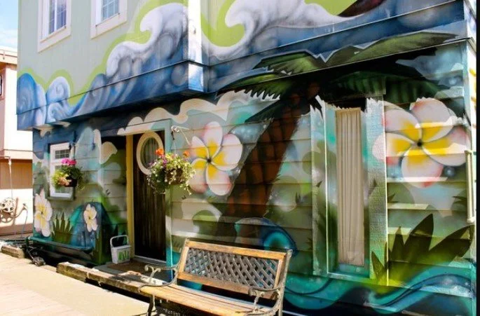 Colorful mural of flowers, a palm tree, and clouds on the exterior wall of a building, with a bench and hanging flower pots in front.