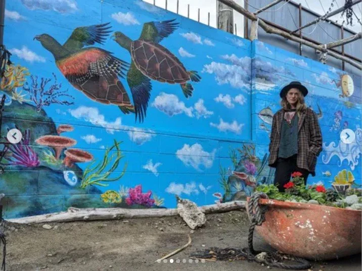 A person standing next to a colorful mural of underwater and sky scene on a wall, featuring flying and swimming sea turtles, vibrant corals, and a bright blue sky with clouds.