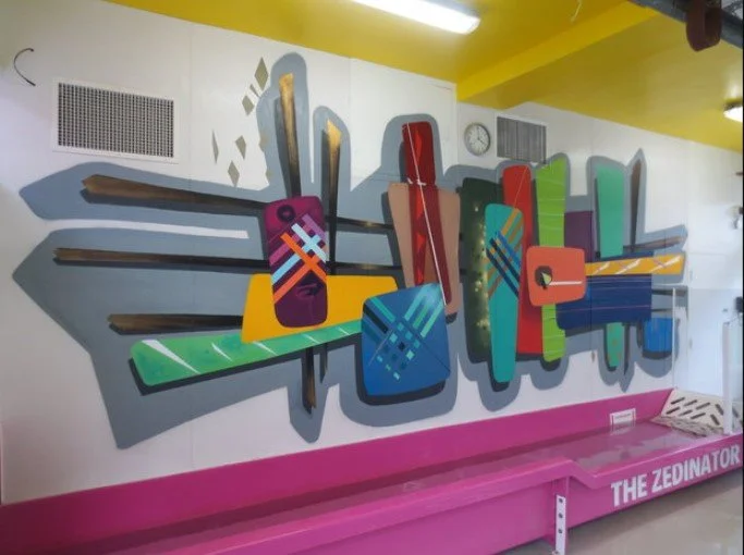 Colorful abstract mural featuring geometric shapes and stylized pencils on a white wall, with a pink base and the words 'The Zedinator' written on it.
