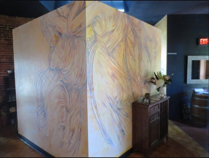 Interior corner of a room with colorful abstract mural on walls, a wooden cabinet with flowers, and a wine barrel on the floor.
