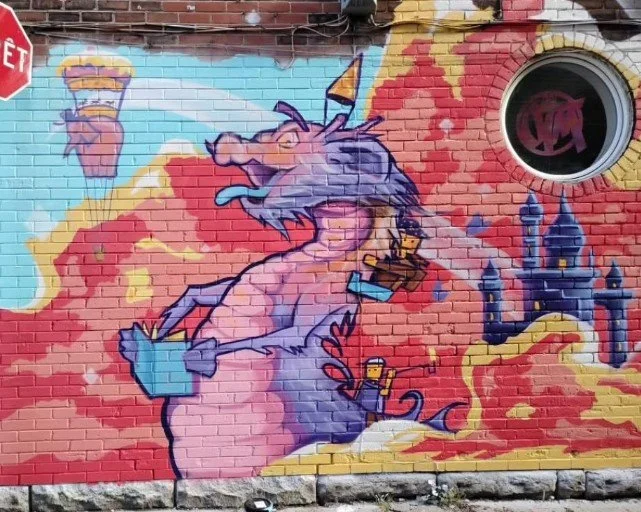 Colorful graffiti of a purple dragon sitting on clouds, holding a paintbrush, near a castle, painted on a brick wall.