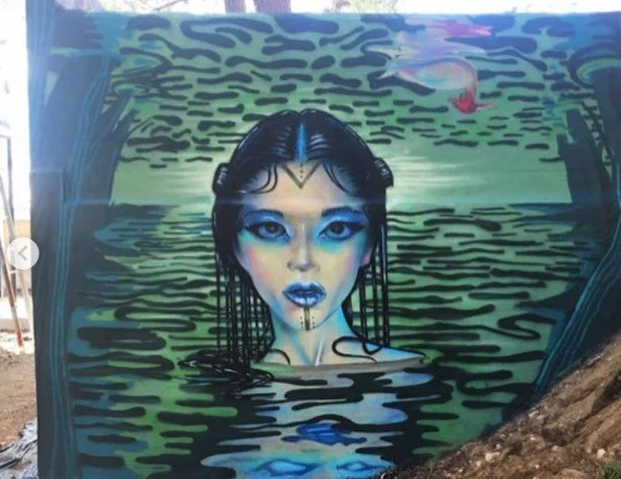 A mural depicting a woman with long dark hair, striking blue makeup, and tribal markings, emerging from a body of water, with a swan flying in the sky in the background., Art, street art, painter, mural, graffiti, spray paint, oil paint, montreal, co