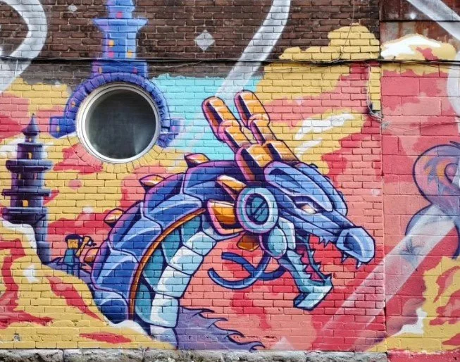 Graffiti mural of a blue dragon with a mechanical appearance, breathing flames on a colorful brick wall.