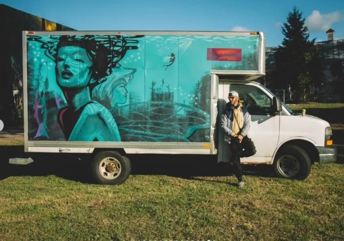 A man standing next to a moving truck with vibrant mural art of a woman's face on the side.