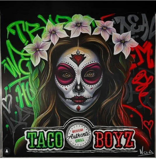 A woman with a sugar skull face paint, wearing a crown of pink and white flowers and neon graffiti in the background, advertising Taco Boyz Authentic Grill.
