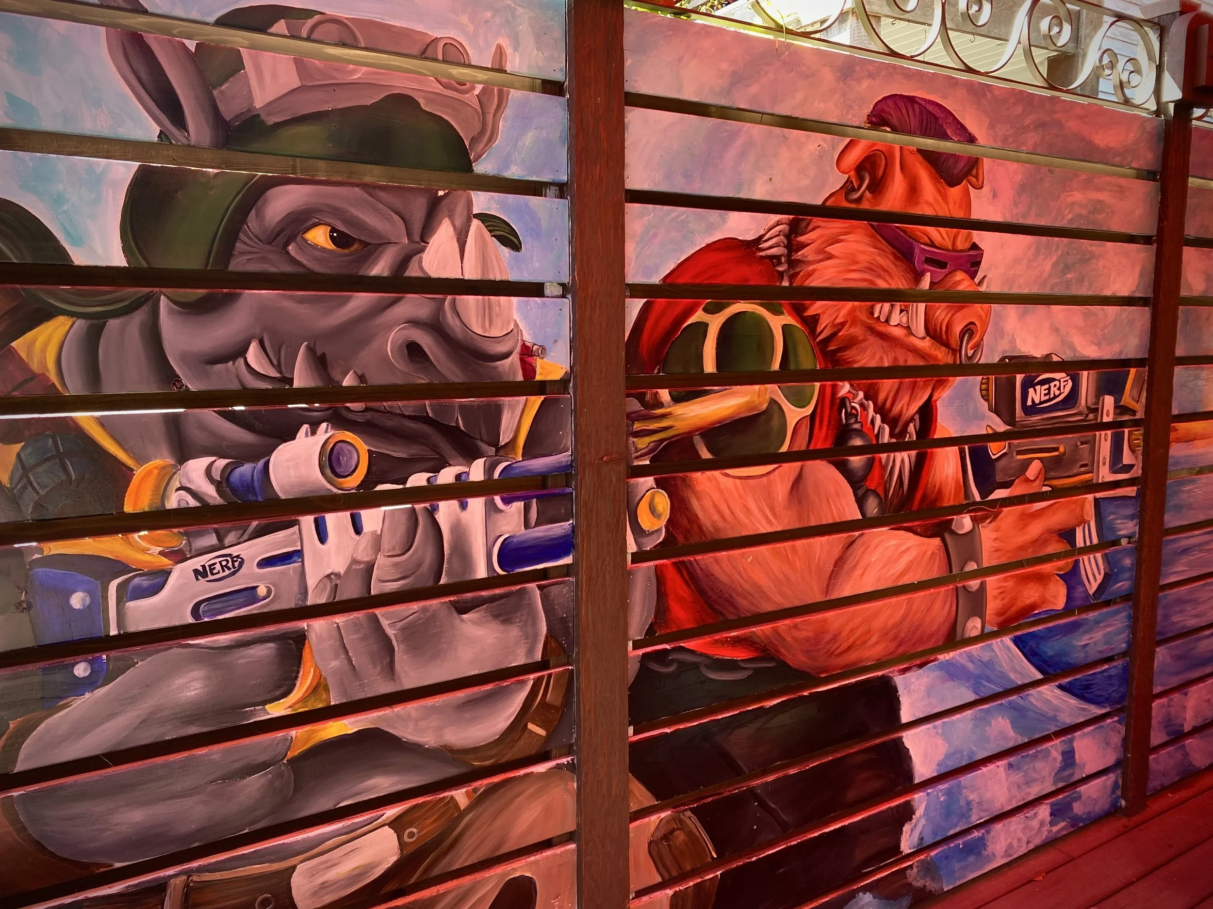 Colorful painting of a rhinoceros and a gorilla, separated by horizontal wooden slats, with a sky background and intricate ironwork at the top.