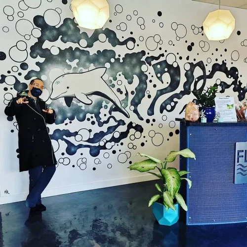 Person wearing a black coat and face mask standing indoors next to a blue counter, with a large wall mural of a dolphin and abstract black and white patterns behind them. There is a green plant in a blue pot in front of the counter.