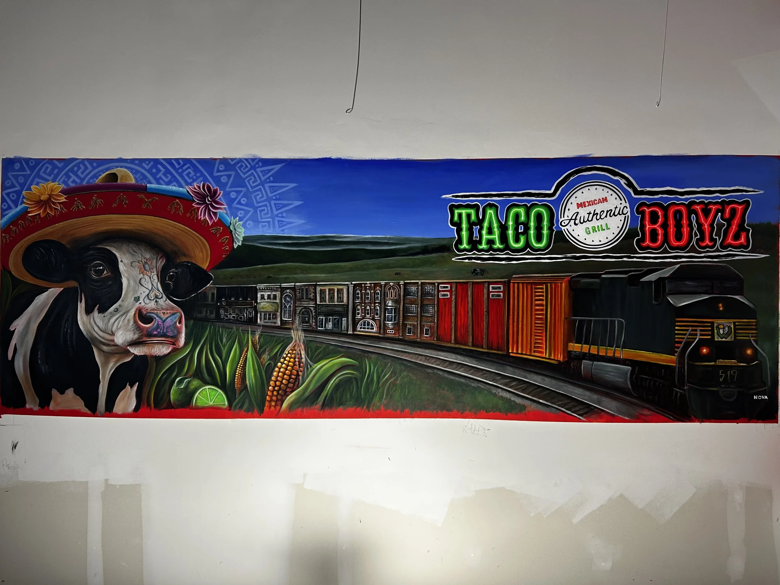Colorful mural featuring a cow wearing a sombrero, with text that says 'Taco Boyz' and includes a landscape scene, train tracks, and a blue sky.