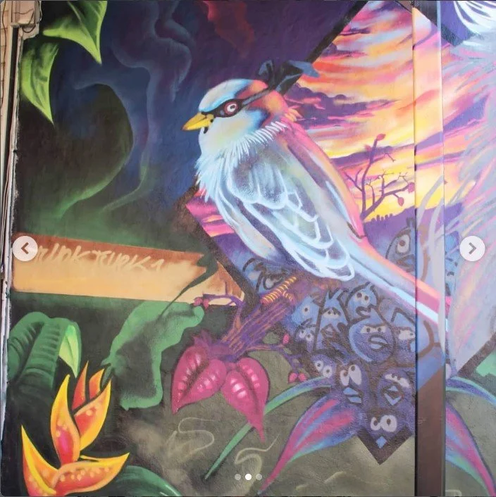 Colorful mural of a bird perched on a branch with vibrant flowers and leaves, set against a sunset sky with a dark silhouette of trees. Art, street art, painter, mural, graffiti, spray paint, oil paint, montreal, commission, Futurism, retro Futurism,