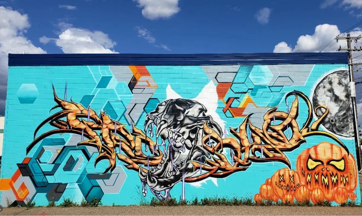 Colorful graffiti mural on a brick wall featuring a dragon, a feline skull, spiders, and jack-o'-lanterns against a bright blue sky with clouds and electrical poles.
