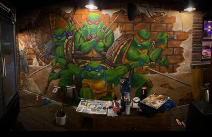 Graffiti art of four Teenage Mutant Ninja Turtles, painted on a brick wall, with art supplies and tools on a table in front.