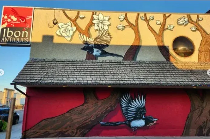 A mural featuring two magpies perched on a tree branch, with a background of stylized tree branches and flowers, and a small circular window on the upper right. The mural also includes the logo for Ibon Antiques in the top left corner.