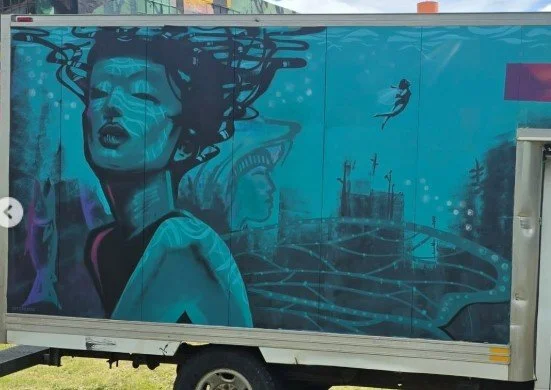 Graffiti mural on a truck trailer shows a stylized woman with flowing black hair, a bridge, and cityscape in shades of blue.