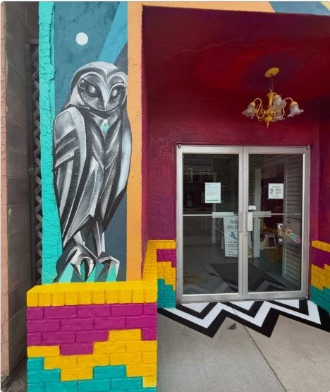 Colorful mural of an owl painted on an exterior wall outside a building with a glass door, a yellow chandelier hanging from the ceiling, and a black and white zigzag patterned sidewalk.