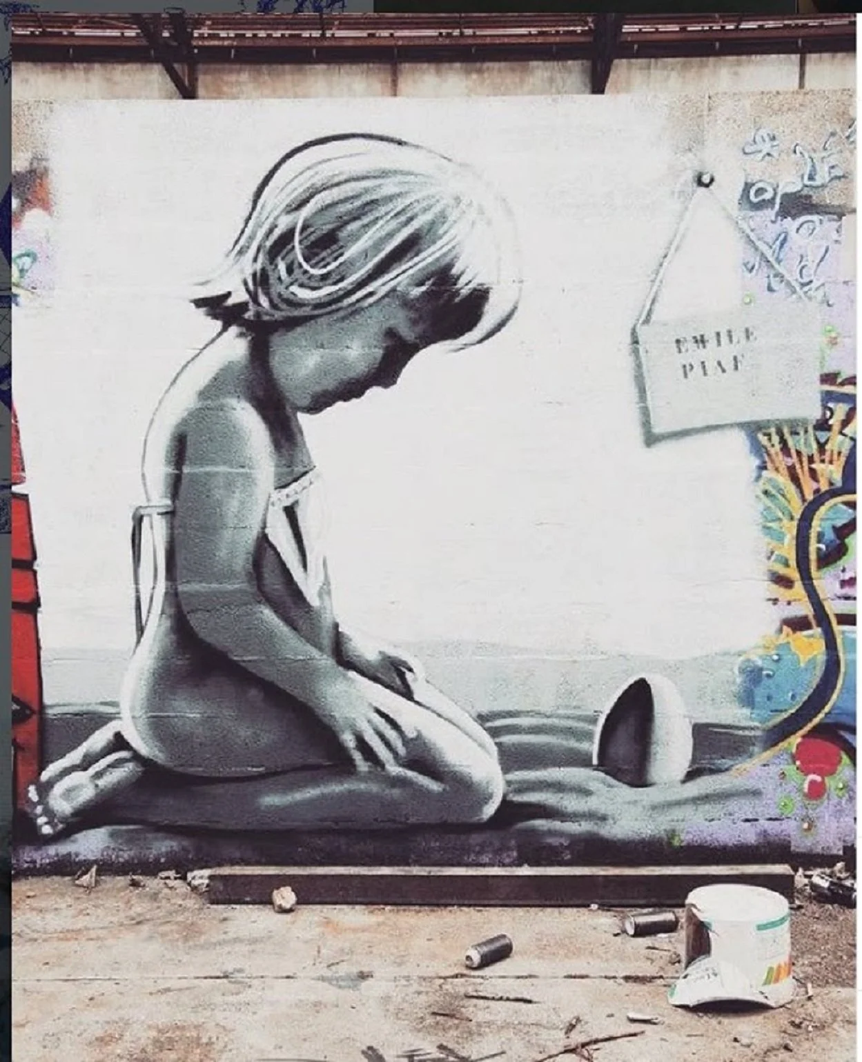 Street art mural of a kneeling girl with short hair, painted in shades of gray, with a small watering can and a sign that reads 'CHILD PLAN' on the wall.