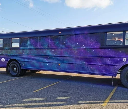 A bus parked in a parking lot with a galaxy-themed paint job featuring purple and blue colors with stars.
