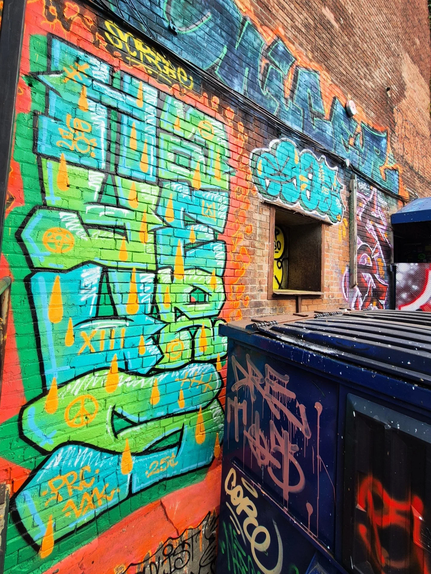 Colorful graffiti art on a brick wall featuring various tags, words, and symbols with a trash bin in the foreground. Art, street art, painter, mural, graffiti, spray paint, oil paint, montreal, commission, Futurism, retro Futurism, cyber punk, punk, 