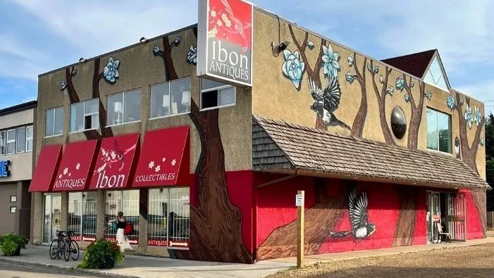 Building with murals of trees, blue flowers, and birds, featuring signs for Ibon Antiques and Collectibles, with red awnings and large glass windows at the storefront.