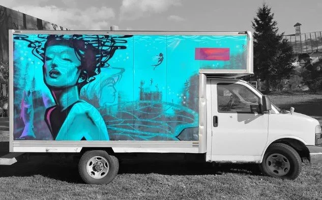 A white delivery truck with a large colorful mural on the side featuring a woman with an elaborate hairstyle, surrounded by abstract blue and purple designs.