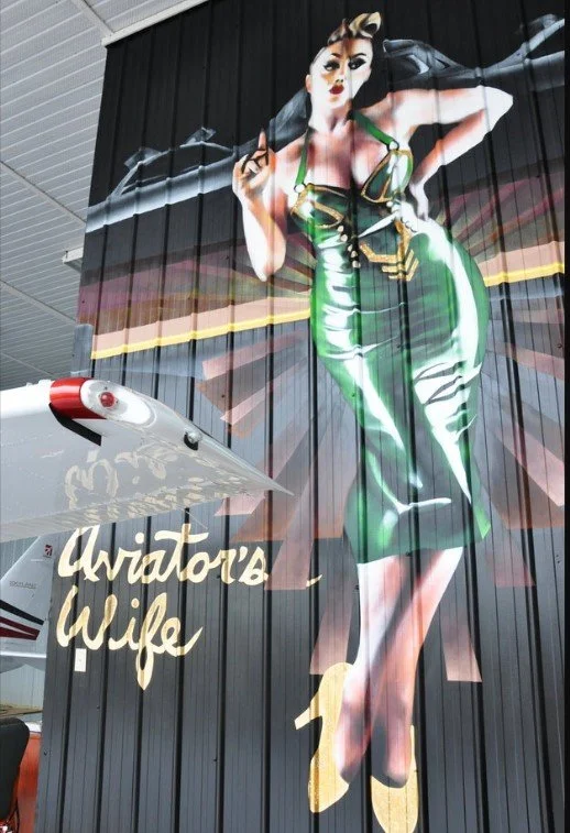 Large mural of a woman in a vintage green dress, holding an umbrella, with handwritten-style text that reads "Aviator's Wife".