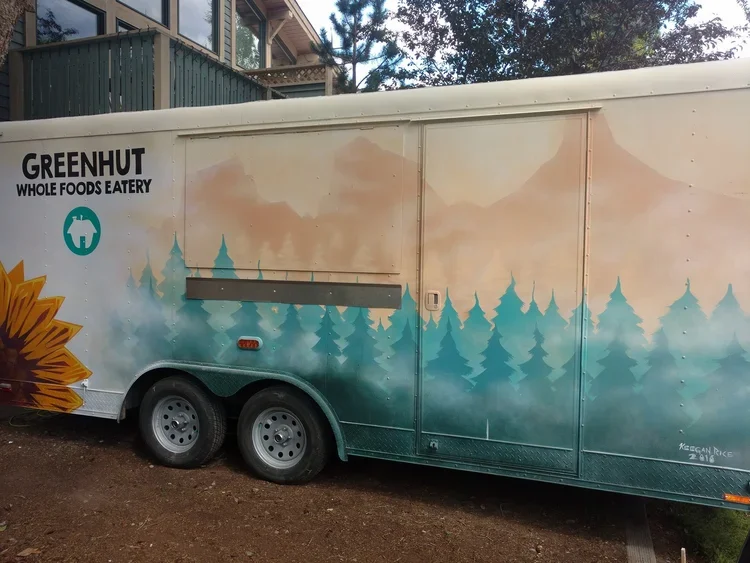 A food truck with a painted outdoor scene of trees and sky, featuring the text 'Greenhut Whole Foods Eatery.'