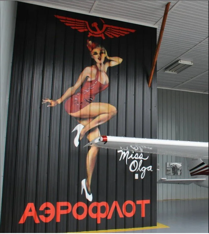 A vintage-style pin-up themed mural of a woman in a red dress and white high heels, with an airplane wing in front of her and a Soviet Union emblem above, on a black wall with the text 'Miss Olga' and Cyrillic writing.
