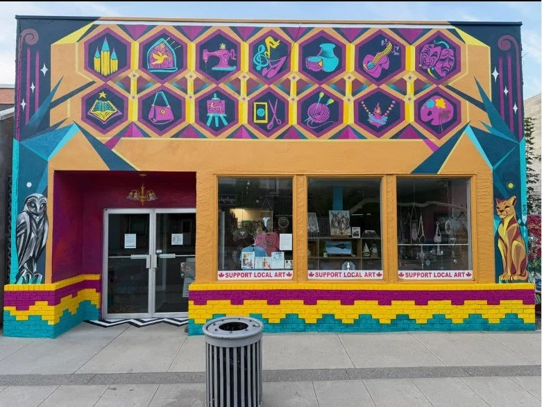 Colorful mural on the building's exterior features geometric and animal illustrations, including an owl on the left and a lioness on the right, with hexagonal icons representing various arts and crafts above the windows. The storefront promotes suppo