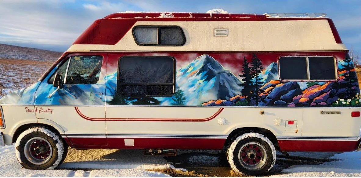 A vintage camper van with a mountain landscape mural painted on its side, parked on snow-covered ground against a partly cloudy sky.