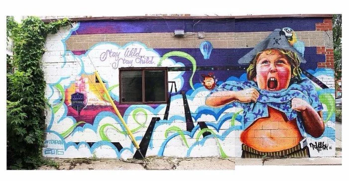 Colorful mural depicting a young boy with a bird on his shoulder, shouting and pulling up his shirt to reveal his stomach, with clouds, a smiling girl, and abstract shapes in the background. Art, street art, painter, mural, graffiti, spray paint, oil