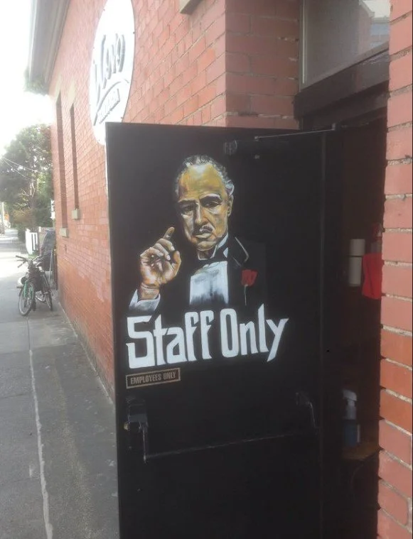A black door features a painted portrait of a man in a tuxedo, pointing a finger. The door has a sign that reads 'Staff Only' and 'Employees Only'. The background shows a brick wall and an outdoor sidewalk with a bicycle.