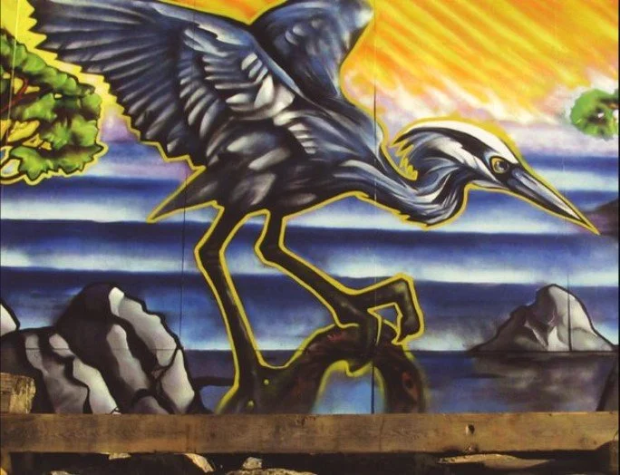 Colorful mural of a large heron bird standing on rocks near water with a sunset sky in the background.