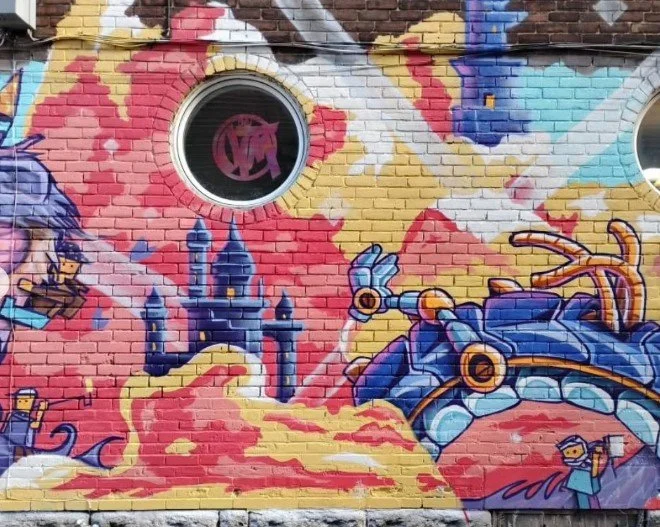 Colorful graffiti mural on brick wall featuring a castle, a robot, and a flying character, with a circular window reflecting the logo 'VM'.