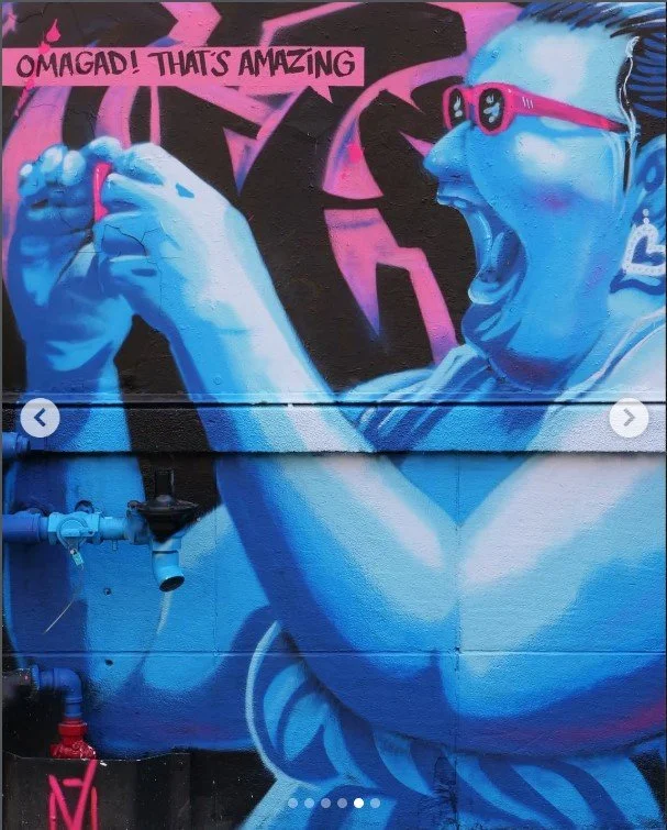 A mural of a woman with blue skin and pink glasses, cheering with her fists raised. The background contains pink and black graffiti, and a speech bubble says, "OMGAD! THAT'S AMAZING.", Art, street art, painter, mural, graffiti, spray paint, oil paint
