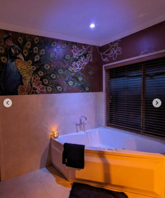 A bathroom with a corner bathtub next to a window with blinds, a peacock painting with colorful feathers on the wall, and a small candle on a ledge near the bathtub, illuminated by a purple ceiling light.