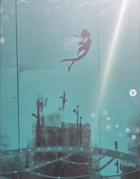 Wall art depicting a mermaid with flowing hair, swimming above a silhouette of underwater plants and a bridge in an aquatic scene.