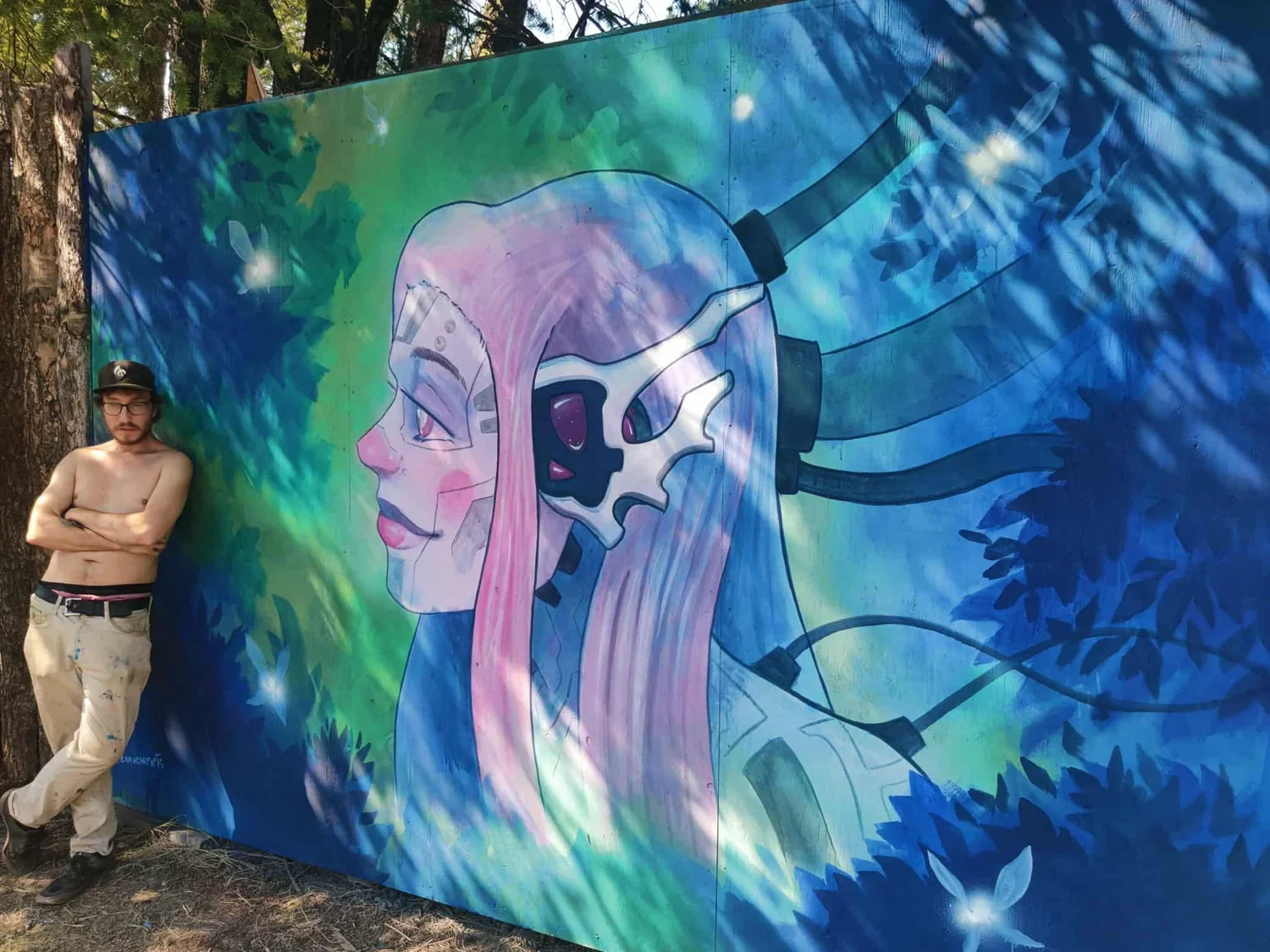 A large colorful mural of a female robot or cyborg with pink hair, wearing a headset, with a decayed skull part on the side of the head. The background has blue and green tones with leafy patterns. A shirtless man with glasses, a black cap, and beige