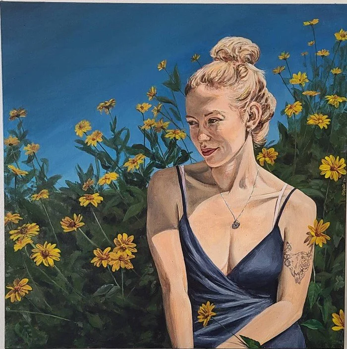 A woman with blonde hair tied up in a bun, wearing a dark blue spaghetti strap dress, sitting among yellow flowers and greenery against a bright blue sky, looking to the side with a slight smile.