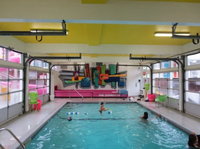 Indoor swimming pool with children swimming and playing, colorful chairs, and a graffiti-style mural on the wall
