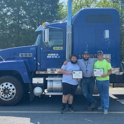 Three men standing in front of a large blue truck, holding certificates, outdoors on a sunny day.