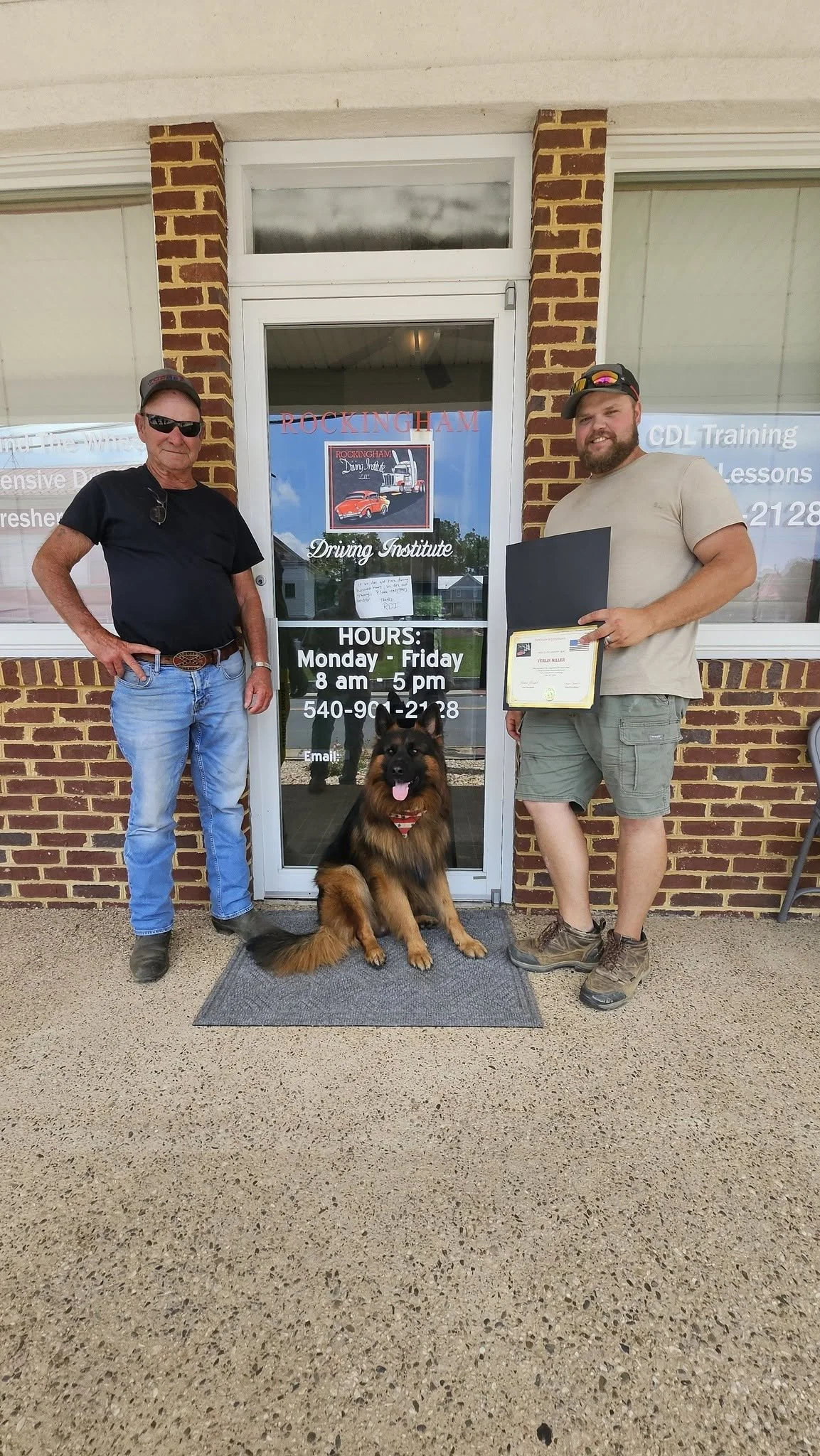Two men and a German Shepherd dog standing in front of a driving school entrance. One man is wearing sunglasses and a black shirt, the other is smiling and holding a certificate. The dog is sitting on a mat, panting, with a red bandana around its neck.